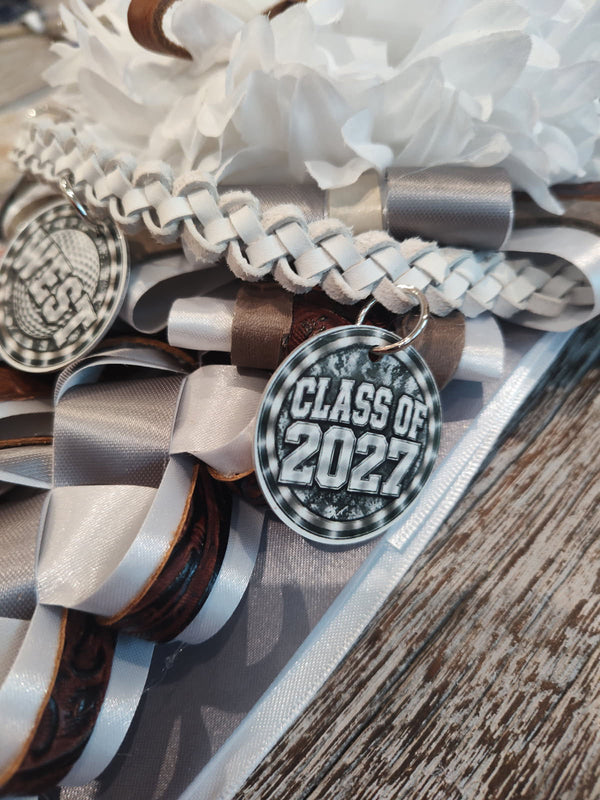 Close-up of a garter with a white braided ribbon, brown and silver loops, and a Class of 2027 charm