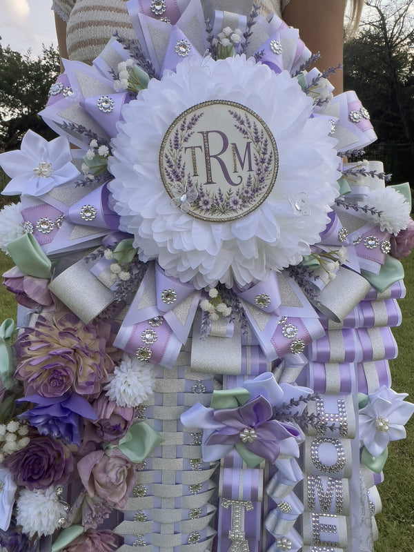 Close-up of a lavender and white homecoming mum with a monogram center, rhinestone loops, and silk flowers