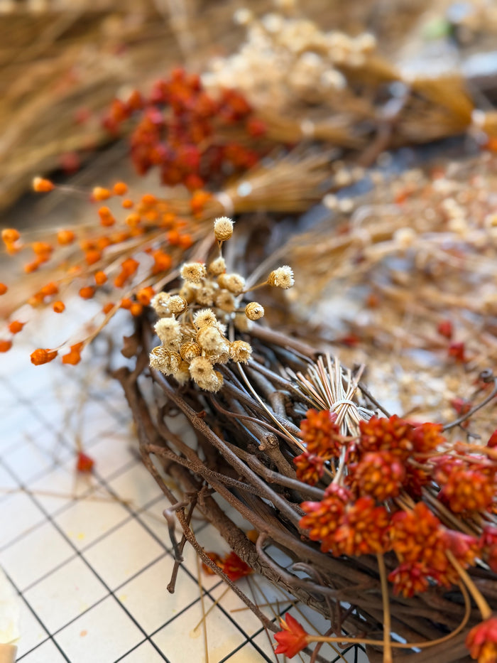 Close-up of a fall wreath in progress on the worktable, with dried orange flowers, baby's breath, and wheat on a twig base