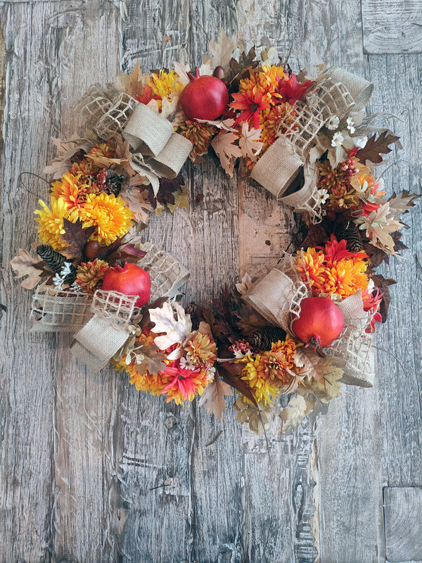 Fall wreath of orange and gold chrysanthemums, red pomegranates, pinecones, and burlap ribbon loops laid flat on rustic wood