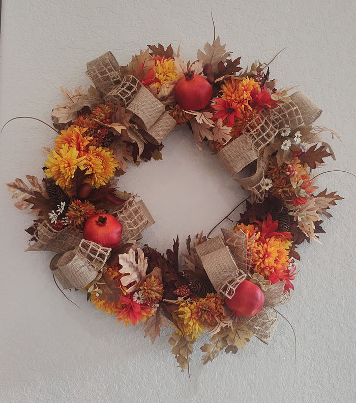 Fall wreath with orange and gold chrysanthemums, red pomegranates, pinecones, autumn leaves, and burlap bows, hanging on a white wall